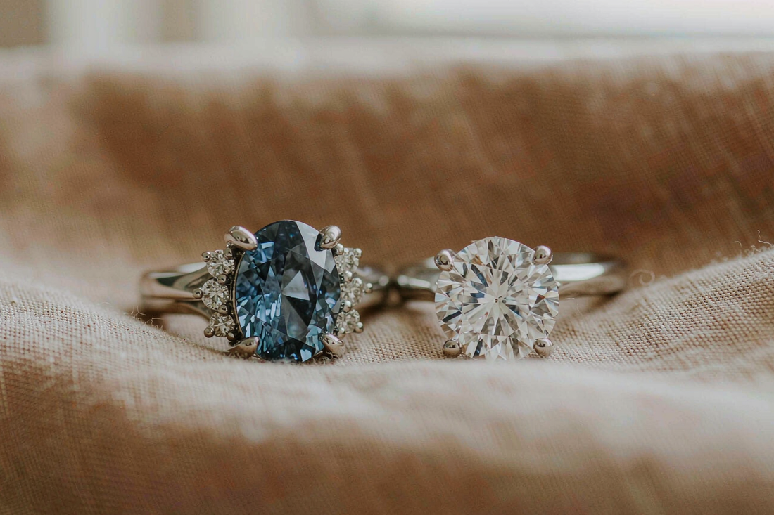 round moissanite engagement ring and oval blue sapphire ring side by side on warm beige linen fabric