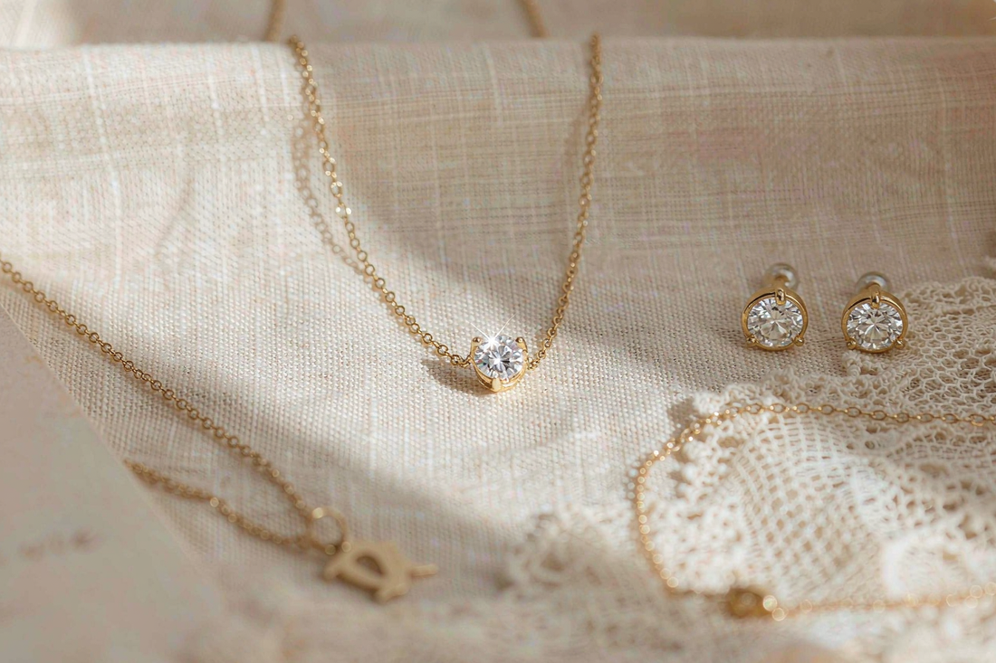 flat lay of mothers day jewelry gift options including moissanite pendant necklace stud earrings and dainty bracelet on cream linen