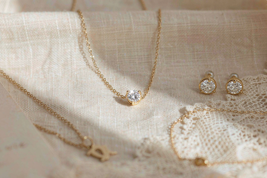 flat lay of mothers day jewelry gift options including moissanite pendant necklace stud earrings and dainty bracelet on cream linen