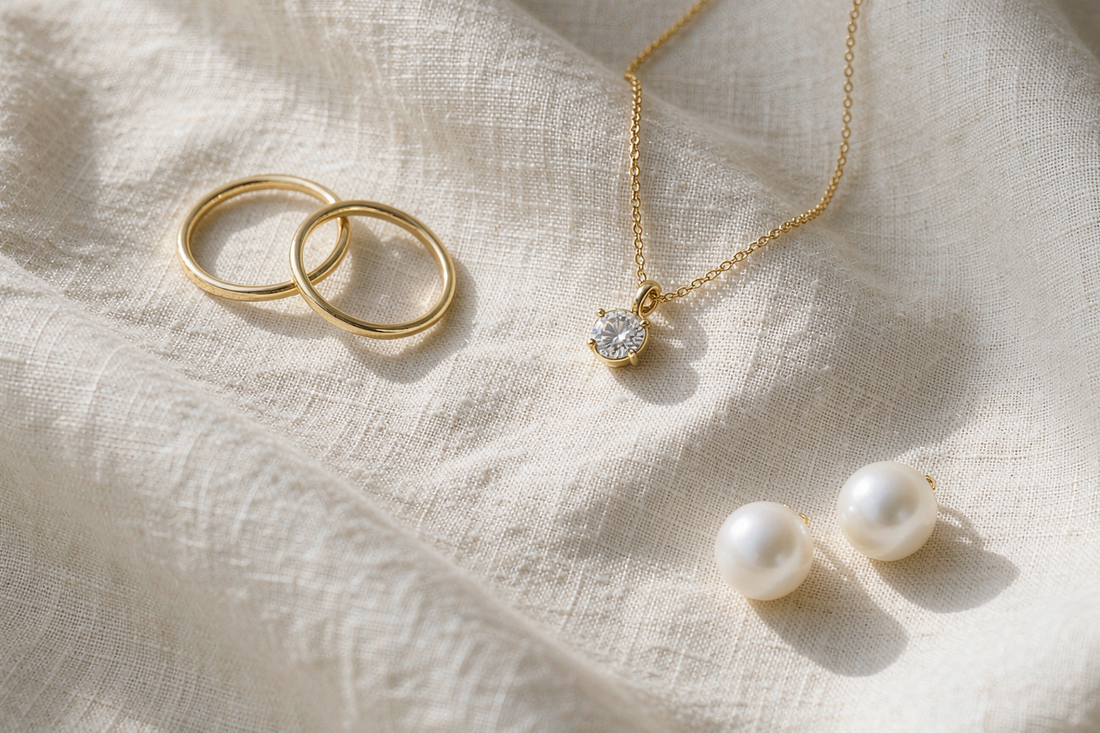 refined quiet luxury jewelry flat lay with thin gold bands pearl studs and small solitaire necklace on cream linen