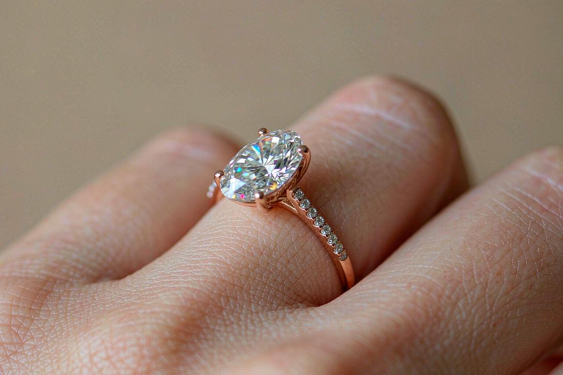 oval moissanite engagement ring on a wider finger with rose gold band on neutral beige background